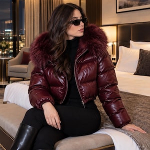 May include: A cropped, shiny burgundy puffer jacket featuring a large, plush faux fur hood in a matching deep red tone. The jacket has a front zip closure, long sleeves with elastic cuffs, and a quilted design, worn over a black turtleneck.