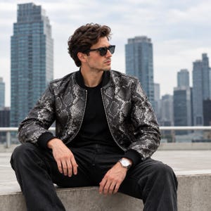 May include: A silver and black snakeskin print bomber jacket with a black ribbed collar, cuffs, and hem. The jacket features a full-length silver zip down the centre, worn over a plain black t-shirt with black jeans and a silver-toned wristwatch.