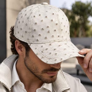 May include: A white baseball cap featuring a textured surface with a repeating pattern of small, circular indentations. The cap has a structured crown, a curved brim, and a small button at the top centre, crafted with a distinct quill-like finish.