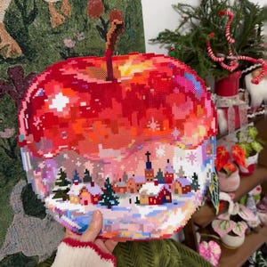 May include: A pixel art apple made of fused plastic beads. The top half is a vibrant red and orange gradient, while the bottom half depicts a snowy winter village scene with small houses, evergreen trees, and a light dusting of white snow.