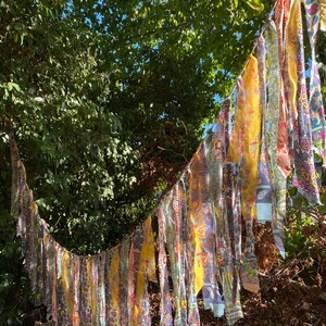 May include: A decorative garland made of numerous hanging fabric strips in various colours and patterns, including yellow, blue, pink, and white. The strips are tied to a central cord, creating a long, flowing, and layered textile display.