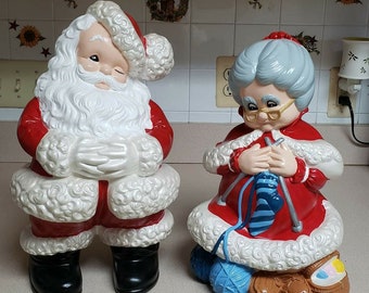 May include: A vintage ceramic Santa Claus and Mrs. Claus figurine set. Santa is wearing a red suit with white trim and a white beard. Mrs. Claus is wearing a red dress with white trim and is knitting a blue sock.