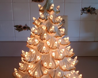 May include: A white ceramic Christmas tree with gold accents and a star on top. The tree is lit with white lights.