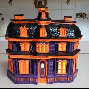 Puede incluir: Una casa de Halloween morada y negra con bordes y ventanas naranjas. La casa tiene un techo negro y una puerta blanca. Las ventanas tienen luces naranjas en el interior.
