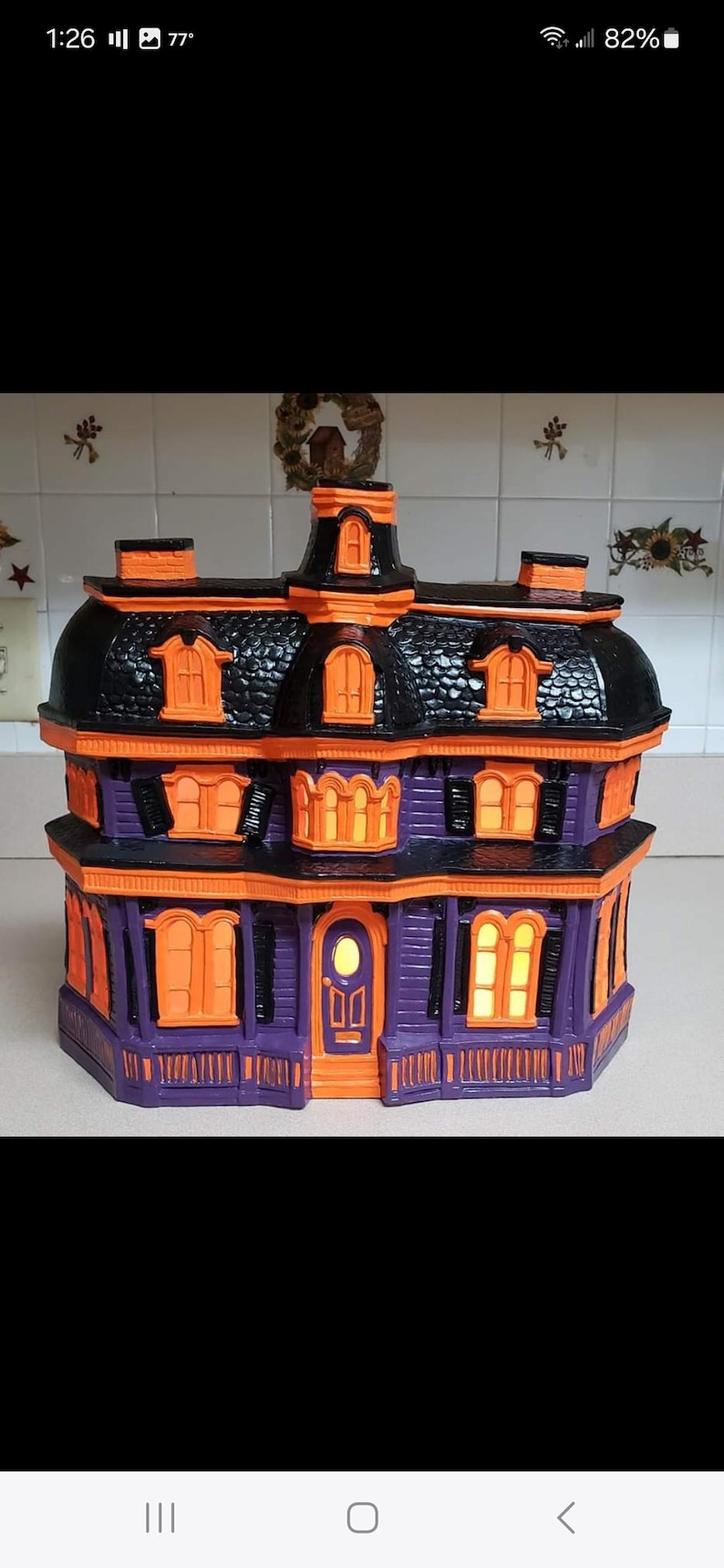 Puede incluir: Una casa de Halloween morada y negra con bordes y ventanas naranjas. La casa tiene un techo negro y una puerta blanca. Las ventanas tienen luces naranjas en el interior.