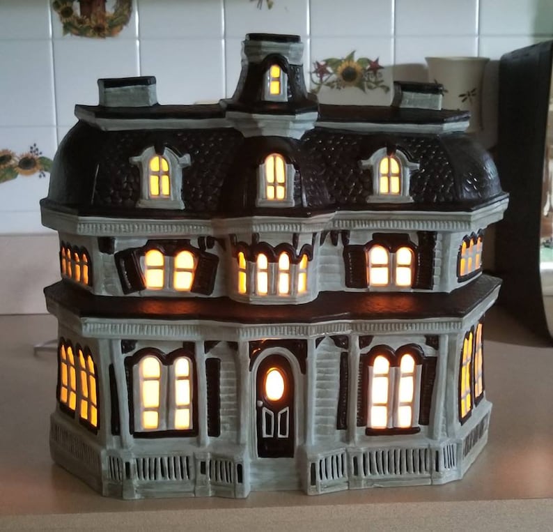 Puede incluir: Una casa encantada de cer&aacute;mica con ventanas iluminadas, un esquema de colores gris y marr&oacute;n y un dise&ntilde;o detallado. La casa tiene un porche delantero con una puerta y un techo con una ventana abuhardillada.