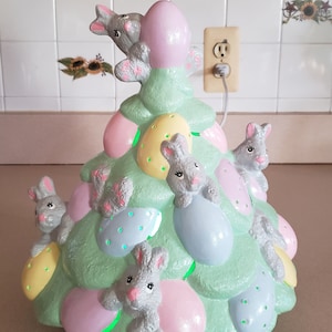 May include: A pastel green ceramic Easter tree with light-up eggs and gray bunny figurines. The tree features pink, yellow, and blue eggs, and is decorated with several small, gray bunnies. The tree is plugged into an electrical outlet.