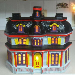 Puede incluir: Una casa encantada de cer&aacute;mica con techo negro, bordes rojos y ventanas amarillas y moradas. La casa tiene un porche con una puerta amarilla y una barandilla blanca.