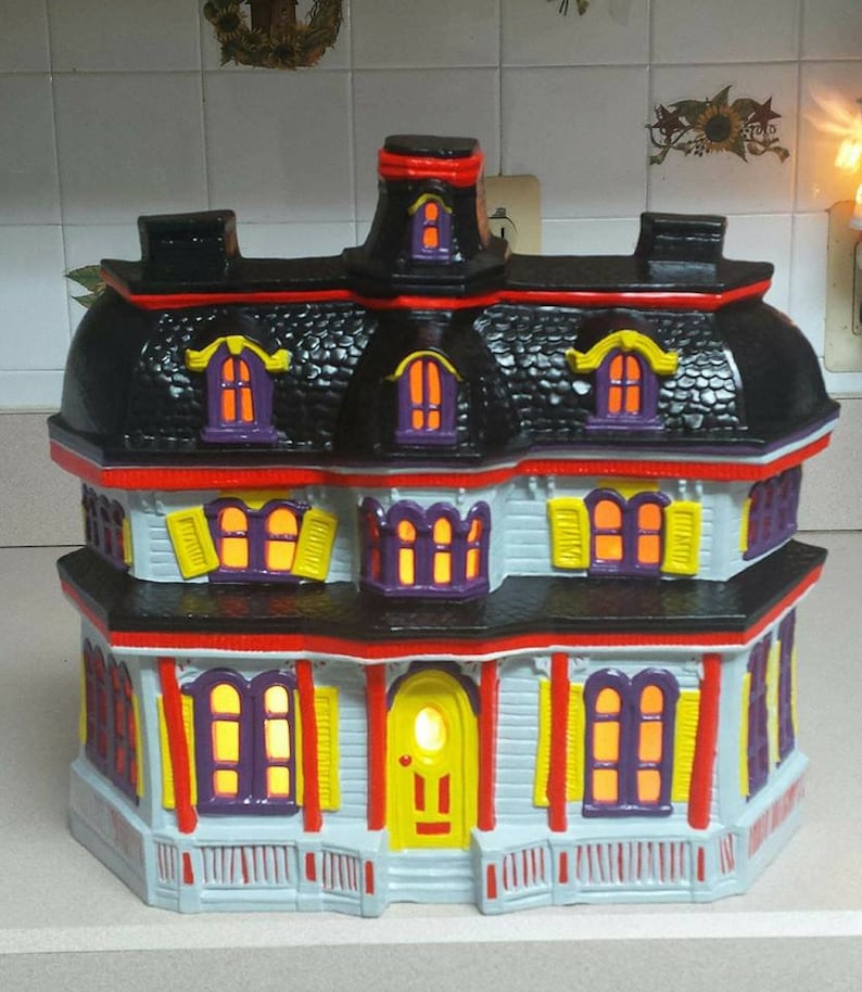 Puede incluir: Una casa encantada de cer&aacute;mica con techo negro, bordes rojos y ventanas amarillas y moradas. La casa tiene un porche con una puerta amarilla y una barandilla blanca.