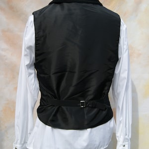 May include: A black velvet-collared vest with a black satin front and a black adjustable strap. The vest is worn over a white dress shirt with ruffled cuffs.