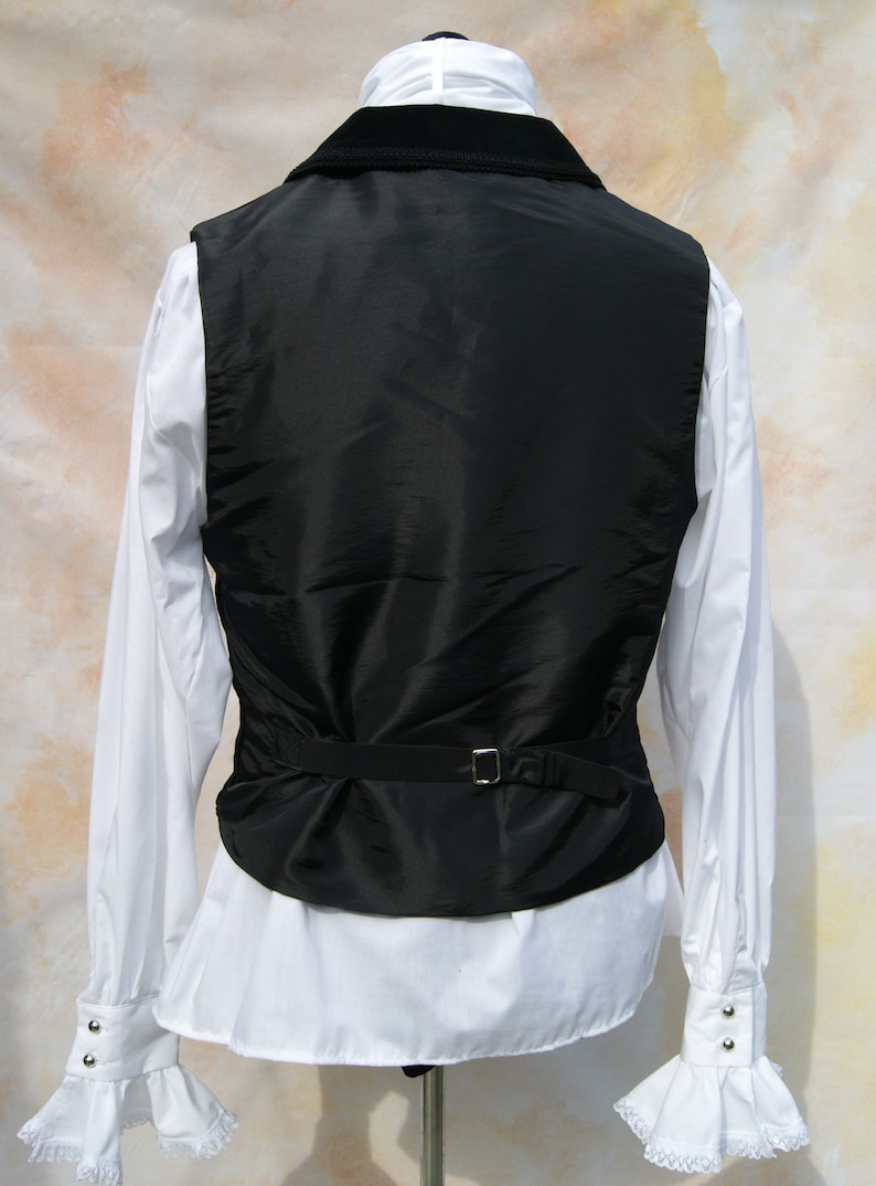 May include: A black velvet-collared vest with a black satin front and a black adjustable strap. The vest is worn over a white dress shirt with ruffled cuffs.
