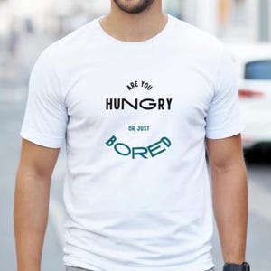 以下が含まれることがあります： 胸元に黒い文字でARE YOU HUNGRY、その下に青緑色の文字でOR JUST BOREDとアーチ状にプリントされた白い半袖Tシャツ。クルーネックで標準的なフィット感のシンプルなデザインの衣類です。
