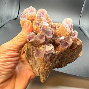 May include: A large cluster of amethyst crystals featuring translucent, light purple, pointed terminations emerging from a textured, tan-coloured rocky base. The mineral specimen displays multiple jagged crystal points with varying degrees of clarity and colour saturation across the formation.