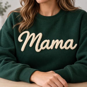 Könnte beinhalten: Ein dunkelgrüner Rundhals-Pullover mit dem Wort Mama in einer erhabenen, strukturierten, cremefarbenen Chenille-Schrift auf der Brust. Das Kleidungsstück hat lange Ärmel mit gerippten Bündchen und einen passenden gerippten Halsausschnitt.