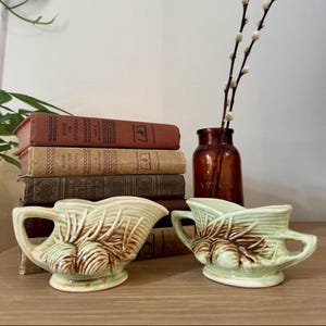 May include: Two pale green ceramic creamers featuring a raised pinecone and needle design with brown accents. Each creamer has a curved handle and a pouring spout. The pieces are positioned in front of a stack of five vintage hardcover books.