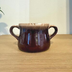 May include: A two-handled ceramic bowl featuring a deep, glossy dark brown glaze on the lower body that transitions into a lighter, creamy beige drip pattern around the upper rim. The vessel has a wide, rounded base and two symmetrical side handles.