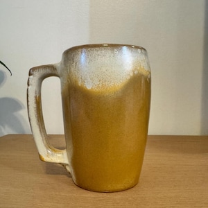 May include: A ceramic mug featuring a two-tone glaze with a smooth, deep mustard yellow finish on the lower portion and a speckled, creamy off-white glaze on the upper rim and handle. The mug has a sturdy, thick handle and a cylindrical body.