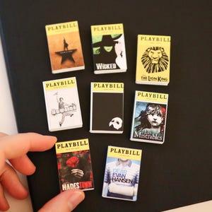 Peut inclure: Une collection de huit répliques rectangulaires miniatures de Playbill présentant diverses couvertures de comédies musicales de Broadway, notamment Hamilton, Wicked, The Lion King, Newsies, The Phantom of the Opera, Les Misérables, Hadestown et Dear Evan Hansen.