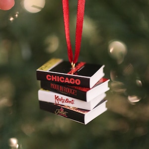 Könnte beinhalten: Ein Miniaturstapel aus vier rechteckigen Buchornamenten, die an einem roten Band hängen. Die Buchrücken zeigen die Titel Chicago, Moulin Rouge!, Kinky Boots und & Juliet in weißer, roter und goldener Schrift auf schwarzem und weißem Grund.