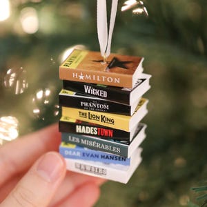 Könnte beinhalten: Ein Stapel Miniatur-Buchanhänger, die an einem weißen Band hängen. Die Buchrücken sind mit Musical-Titeln beschriftet: Hamilton, Wicked, The Phantom of the Opera, The Lion King, Hadestown, Les Misérables, Dear Evan Hansen und Newsies.