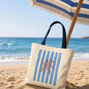 Puede incluir: Un bolso de tela color crema con asas azul marino oscuro, que presenta un patrón de rayas verticales azul claro y un gráfico central de una estrella de mar naranja con el texto "beach please" impreso en una pequeña fuente cursiva azul.