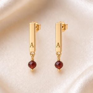 Può includere: Un paio di orecchini pendenti color oro con una barra rettangolare verticale incisa con la lettera A. Ogni barra è fissata a una piccola perla di pietra preziosa rosso scuro sfaccettata che pende dalla parte inferiore della barra.