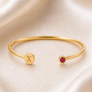 以下が含まれることがあります： 片方の端に文字「X」が刻印された円形のチャーム、もう一方の端にゴールドトーンのベゼルにセットされた小さな丸い深紅の宝石があしらわれた、ゴールドトーンのオープンカフブレスレット。シンプルでバランスの取れたデザインです。