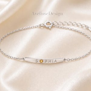 Puede incluir: Una pulsera de cadena plateada con una barra de metal curva grabada con el nombre SOPHIA, donde la letra O se sustituye por una pequeña piedra preciosa redonda de color ámbar. La pulsera incluye un cierre de resorte y una cadena extensora.