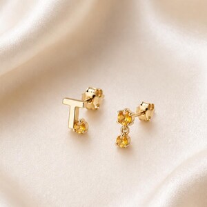 Peut inclure: Une paire de boucles d'oreilles à tige en or, composée d'une lettre T sur l'une et de deux petites pierres précieuses jaunes rondes sur l'autre. Chaque boucle d'oreille présente une finition dorée polie et un fermoir à tige sécurisé.
