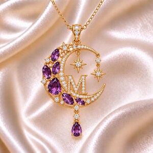 Puede incluir: Un collar con colgante dorado en forma de media luna adornado con gemas moradas de corte marquesa y pequeñas piedras transparentes. La letra M está centrada dentro de la luna, acompañada por dos detalles en forma de estrella y una piedra morada en forma de pera.