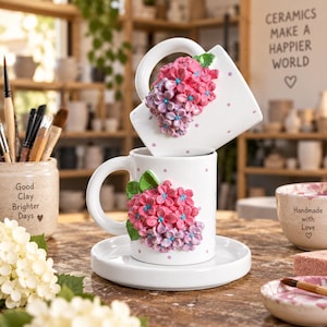 May include: Two white ceramic mugs with pink polka dots and three-dimensional sculpted pink and purple hydrangea flower clusters. One mug rests on a matching white saucer, while the other is balanced on top. Each mug features a curved handle.