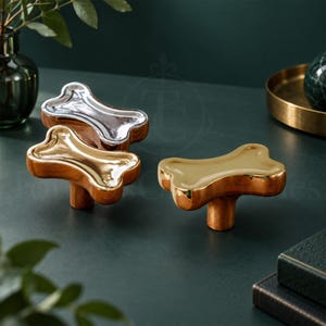 May include: Three bone-shaped cabinet knobs with a smooth, reflective metallic finish. One knob is silver-toned, while the other two are gold-toned. Each knob features a central cylindrical base for attachment, showcasing a polished, shiny surface.