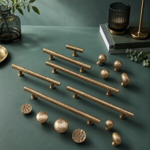 Op de afbeelding: Een set messingkleurige kastbeslagstukken met een gehamerde textuur. De collectie bevat diverse lange, cilindrische stanggrepen met twee bevestigingspunten en meerdere ronde, bolvormige knoppen in verschillende formaten met een consistente metaalafwerking.