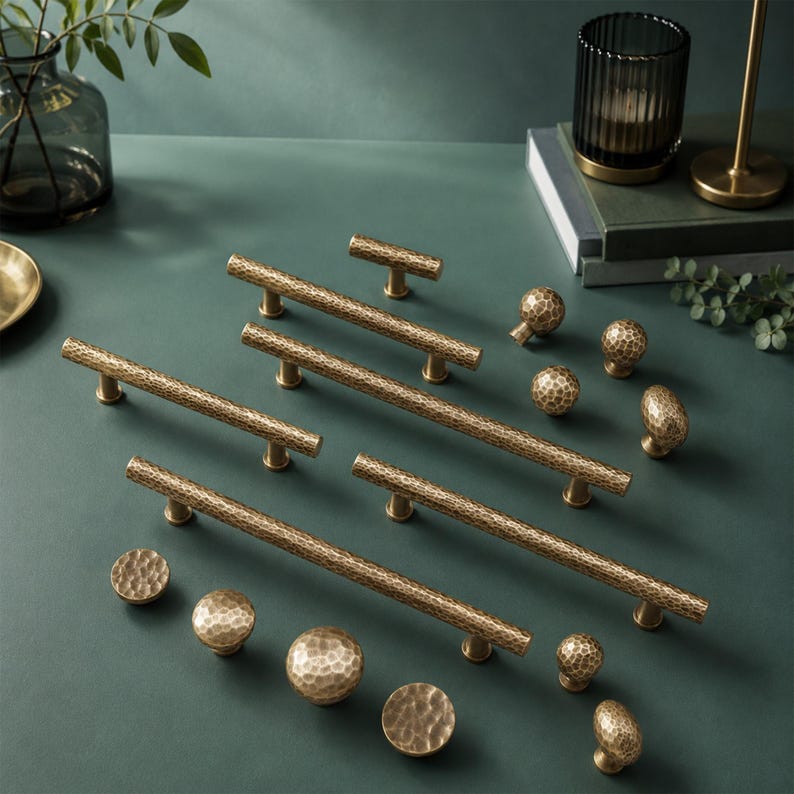 Op de afbeelding: Een set messingkleurige kastbeslagstukken met een gehamerde textuur. De collectie bevat diverse lange, cilindrische stanggrepen met twee bevestigingspunten en meerdere ronde, bolvormige knoppen in verschillende formaten met een consistente metaalafwerking.