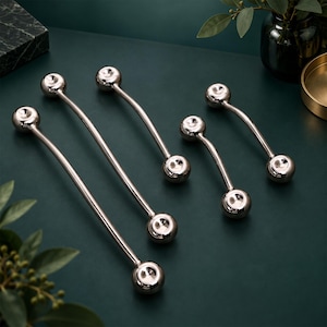 May include: A set of four polished silver-toned metal cabinet pulls, each featuring a curved bar design with spherical ends. The hardware pieces vary in length, showcasing a smooth, reflective finish that highlights the rounded, ball-shaped terminals.