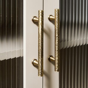 Op de afbeelding: Twee verticale kastgrepen met een cilindrische vorm en een gehamerde messing afwerking. Elke handgreep is met twee ronde voetjes bevestigd aan een lichtgekleurde kastdeur. Het oppervlak van de metalen grepen heeft een textuur met putjes.