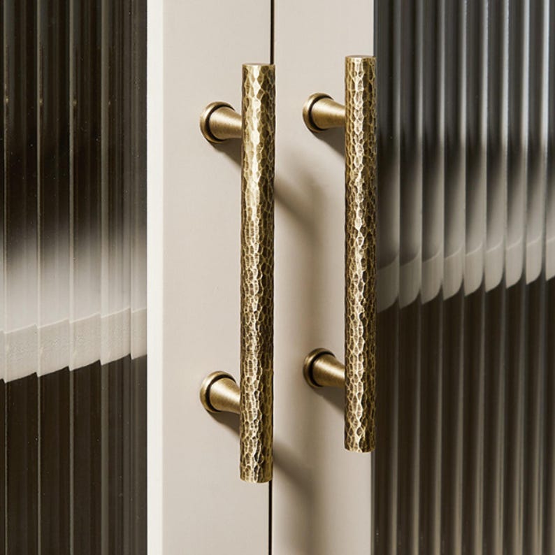 Op de afbeelding: Twee verticale kastgrepen met een cilindrische vorm en een gehamerde messing afwerking. Elke handgreep is met twee ronde voetjes bevestigd aan een lichtgekleurde kastdeur. Het oppervlak van de metalen grepen heeft een textuur met putjes.