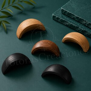 Peut inclure: Cinq poignées de tiroir en bois en forme de croissant disposées en groupe. Deux poignées sont brun clair, une est brun moyen et deux sont noires. Chaque pièce présente une texture de grain de bois visible et un design incurvé et lisse.