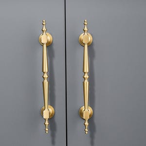 May include: Two vertical brass cabinet pulls with a brushed gold finish. Each handle features a central tapered rod with decorative finials at the top and bottom, secured to the surface by two circular mounting plates with square-shaped bases.