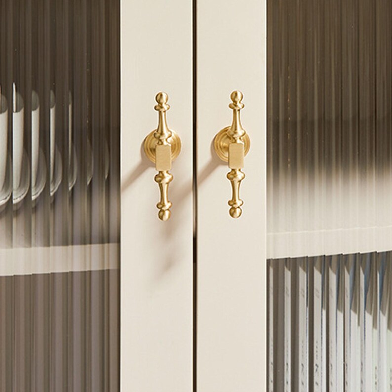 May include: A pair of vertical brass cabinet handles with a decorative spindle shape mounted on a cream-colored cabinet frame. The cabinet features fluted glass panels that partially obscure the contents stored inside the unit behind the doors.