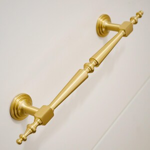 May include: A brushed gold metal cabinet pull handle featuring a long, tapered central bar with decorative spindle-like ends. The handle is mounted on two circular bases and includes small, finial-style points extending from each end of the bar.