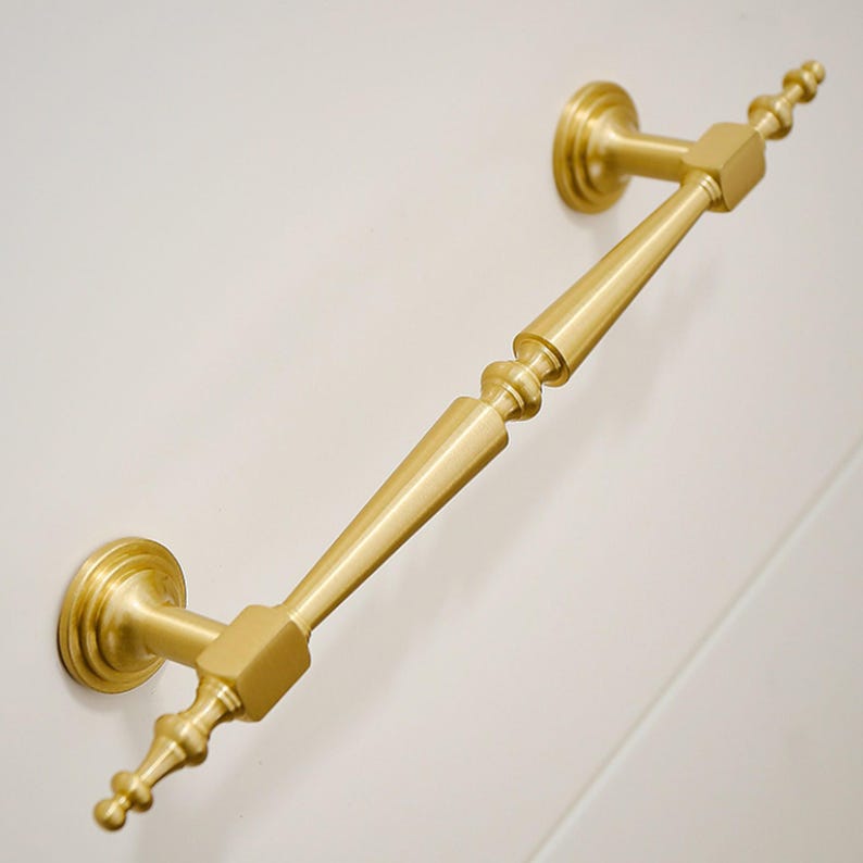May include: A brushed gold metal cabinet pull handle featuring a long, tapered central bar with decorative spindle-like ends. The handle is mounted on two circular bases and includes small, finial-style points extending from each end of the bar.