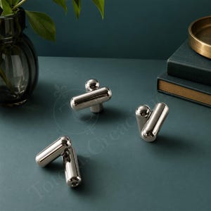 Puede incluir: Tres pomos de metal plateado pulido para muebles, cada uno con un diseño de barra cilíndrica con un extremo redondeado y un pequeño acento esférico. Los herrajes tienen un acabado liso y reflectante, con una construcción sólida y moderna.