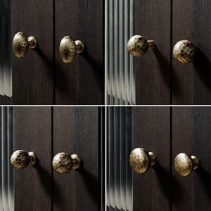 Op de afbeelding: Een collectie van acht ronde kastknoppen met een gehamerd, getextureerd oppervlak in een matte messing afwerking. Elke knop is gemonteerd op een donkerhouten paneel en toont een consistente ronde vorm en een metalen, gedeukt uiterlijk.
