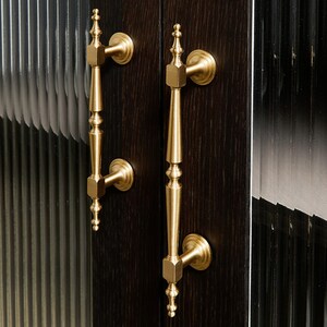 May include: Two vertical brass door handles with a slender, tapered design and decorative finials at the top and bottom. The handles are mounted onto a dark brown, wood-grained door panel positioned next to vertical, fluted glass panels.