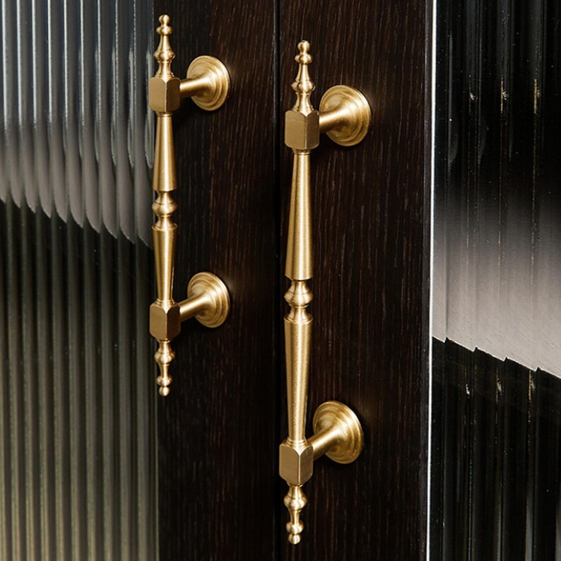 May include: Two vertical brass door handles with a slender, tapered design and decorative finials at the top and bottom. The handles are mounted onto a dark brown, wood-grained door panel positioned next to vertical, fluted glass panels.
