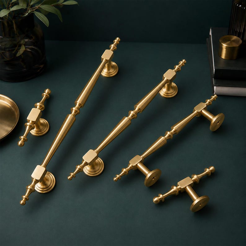 May include: A collection of five gold-toned metal cabinet pulls and knobs of varying lengths. Each piece features a central bar with square accents, tapered ends, and circular mounting bases. The hardware has a smooth, polished finish throughout.