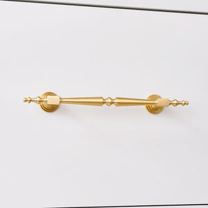 May include: A horizontal drawer pull handle with a brushed gold finish. The handle features a central tapered bar with decorative rounded finials at each end and two square mounting points that connect to circular bases attached to the surface.
