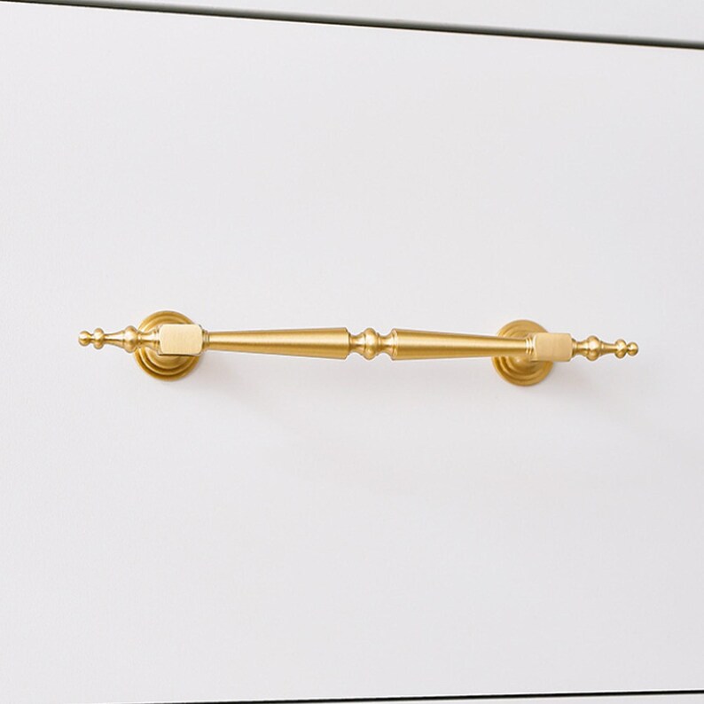 May include: A horizontal drawer pull handle with a brushed gold finish. The handle features a central tapered bar with decorative rounded finials at each end and two square mounting points that connect to circular bases attached to the surface.