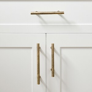 Op de afbeelding: Drie cilindervormige kastgrepen met een getextureerde, gehamerde gouden afwerking bevestigd op witte kastdeuren. E&eacute;n horizontale greep is gecentreerd op de bovenste lade, terwijl twee verticale grepen op de onderste kastdeuren zijn gemonteerd.